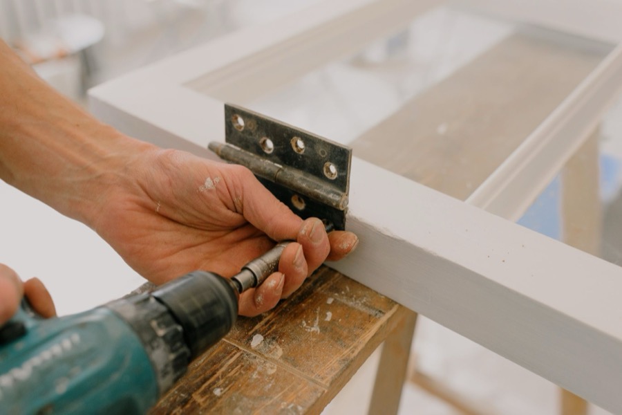 Hands demonstrating the repair of a drywall screw pop, applying joint compound over the re-secured fastener with a putty knife.